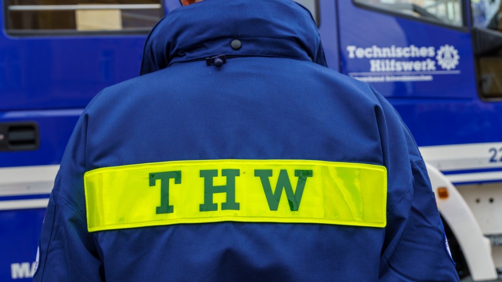 Foto: Ehrenamtlicher Helfer vom Technisches Hilfswerk steht in Uniform vor einem Einsatzfahrzeug des THW 