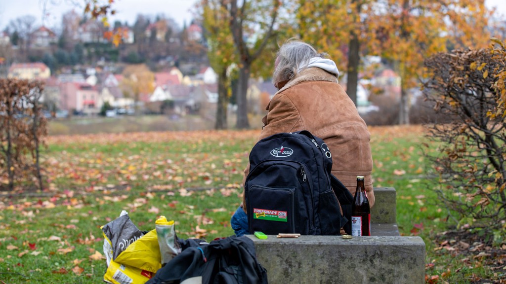 Ein älterer Obdachloser sitzt mit seinem Hab und Gut auf einer Parkbank eines Dorfes