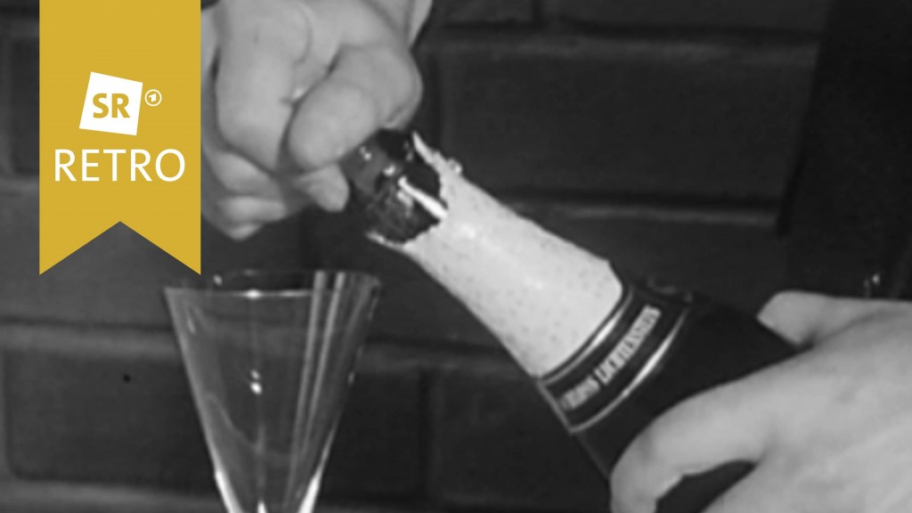 Hand hält Sektflasche. Sektglas steht auf dem Tisch.