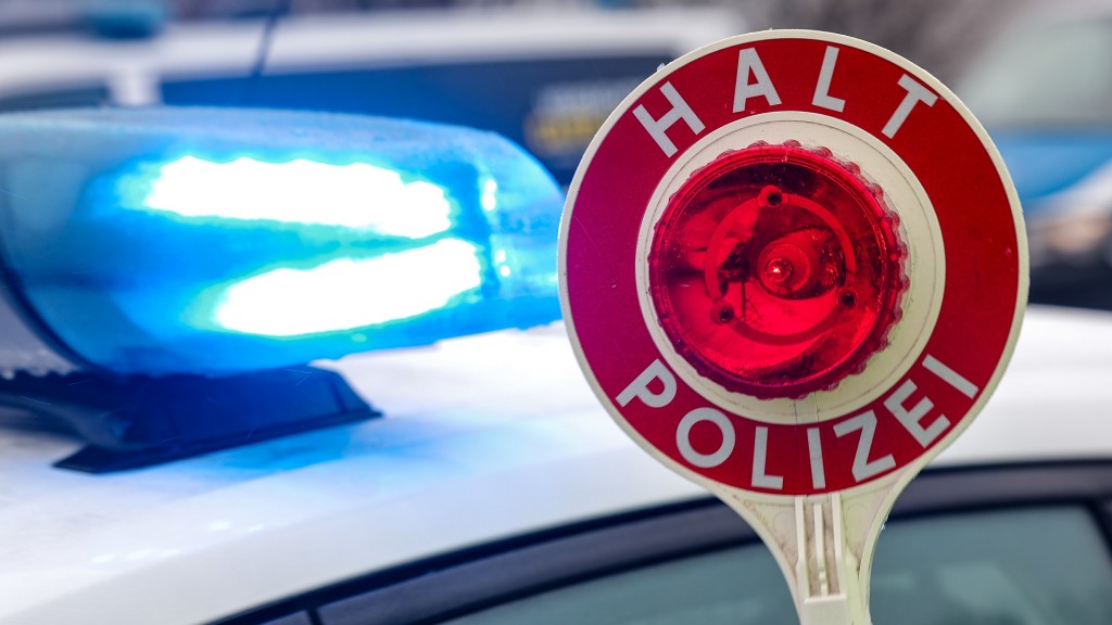 Ein Polizist hält eine Polizeikelle für eine Kontrolle. Im Hintergrund das Blaulicht auf einem Polizeiauto. 