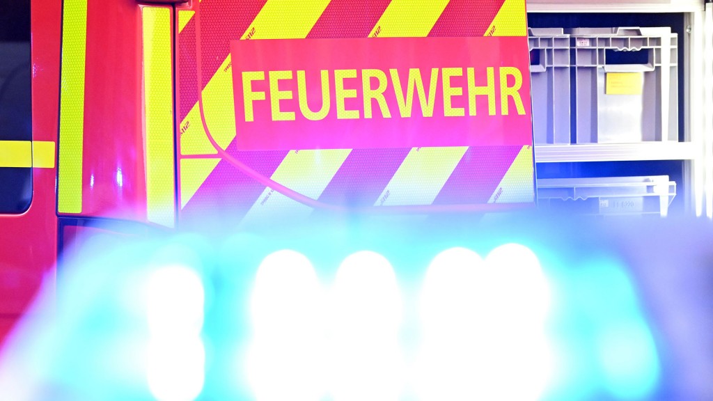 Foto: Ein Fahrzeug der Feuerwehr steht bei einem Einsatz auf einer Straße, während im Vordergrund das Blaulicht eines Polizeifahrzeugs zu sehen ist