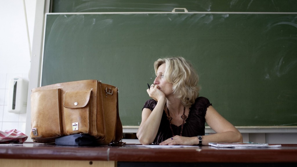 Eine Lehrerin sitzt nachdenklich am Pult vor einer Schultafel.